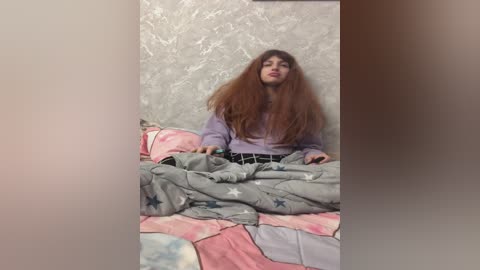 Video of a young woman with long, voluminous brown hair, wearing a lavender sweater and gray pants, lying on a bed with a pink and gray quilt, against a textured silver wall.