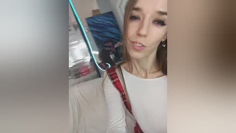 Video of a young, fair-skinned woman with straight brown hair, wearing a white top and red plaid jacket, sitting in a modern, dimly-lit room with a TV and shelves in the background.