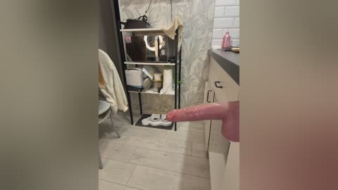 Video of a cluttered bathroom with light-colored tiles and a partially visible erect, circumcised penis being held by a hand. Background shows a small shelf with toiletries and a white chair.