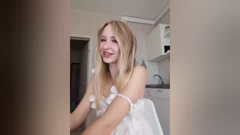 Video of a smiling, blonde woman in a white dress with fluffy white trim, seated in a modern kitchen with white cabinets and a microwave.