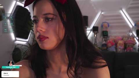 Video of a woman with long dark hair, wearing red devil horns, red lipstick, and a black top, sitting in a futuristic spaceship with hexagonal windows and a toy-filled background.
