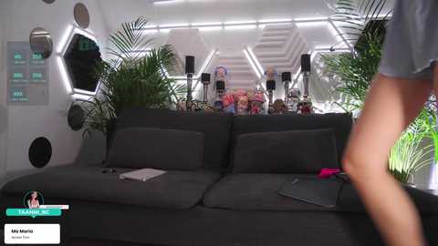 Video of a futuristic room with a grey couch, green plants, and a person in a grey shirt standing. Background features a white, hexagonal wall with monitors displaying green text.