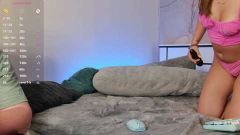 Video of a woman in pink lingerie, kneeling on a gray bed, holding a remote, in a dimly lit room.