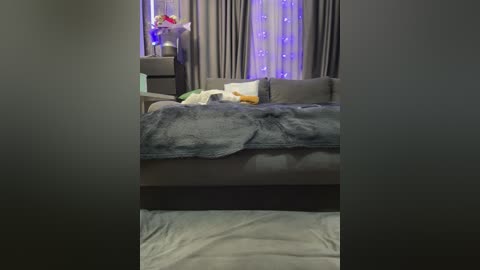 Video of a modern, dimly lit bedroom with a gray sofa, gray throw blanket, white pillows, and a gray carpet. The background features sheer curtains with purple LED lights, adding a soft glow.