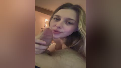 A video showing a young woman with light skin and long blonde hair, partially topless, holding a hard, circumcised penis. The background is dimly lit with beige walls.