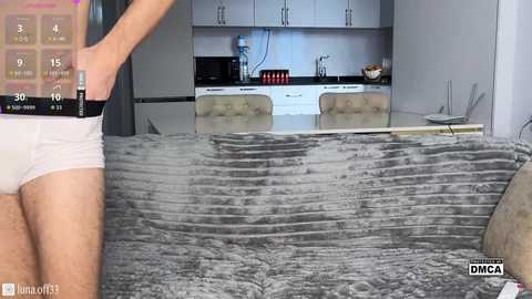 Video of a man in white underwear, standing in a modern kitchen with white cabinets and a gray textured sofa.