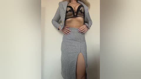 Video of a woman with long blonde hair, wearing a black lace bra and a grey checkered skirt with a high slit, standing confidently against a plain beige wall.