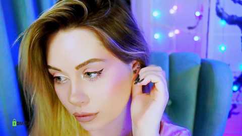 Video of a light-skinned woman with long, blonde hair, wearing black eyeliner, adjusting her earring, in a room with blue and purple lighting, a green chair, and string lights in the background.
