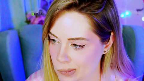 Video of a fair-skinned woman with long blonde hair and blue eyes, wearing subtle makeup, including winged eyeliner, sitting indoors with a blue sofa in the background.