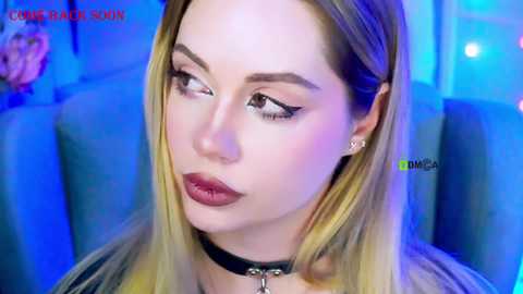 Video of a young woman with fair skin, long blonde hair, and sharp eyebrows, wearing black eyeliner, mascara, and dark lipstick. She has a black choker necklace. Background shows blue lighting and a blurred chair.
