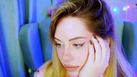 A video of a blonde woman with fair skin, blue eyes, and long hair, resting her head on her hand, gazing thoughtfully. The background features blue curtains and soft, blurred lights.