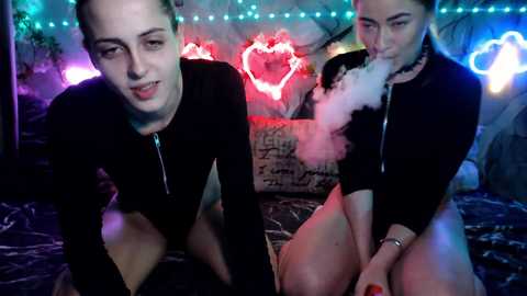 Video of two women with pale skin and blonde hair, both wearing black long-sleeve tops and no bottoms, sitting on a bed with a dark blanket, surrounded by neon lights and a heart-shaped glowing sign.