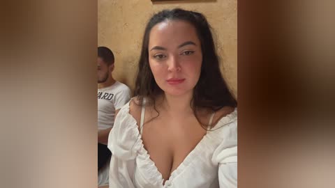 A video of a woman with long dark hair, fair skin, and a curvy physique, wearing a low-cut white blouse, standing in a dimly lit room with a beige wall and another man partially visible in the background.