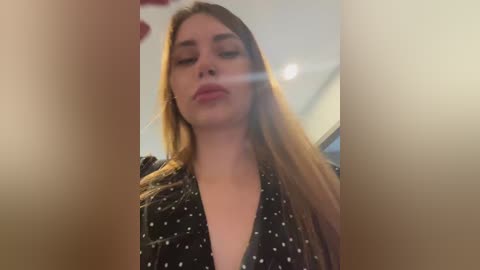 A close-up video of a Caucasian woman with long, straight, light brown hair, wearing a black polka dot blouse. She has fair skin and is looking downward with a slight smile. The background is blurred, suggesting an indoor setting.