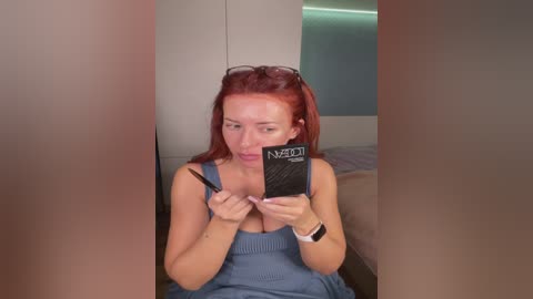 Video of a young, fair-skinned woman with long, red hair, wearing a blue tank top and black sunglasses, taking a selfie in a mirror.