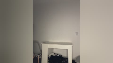 Video of a small, dimly lit room with a white table and two chairs against a plain, light gray wall. The room is cluttered with clothes and personal items on the floor.