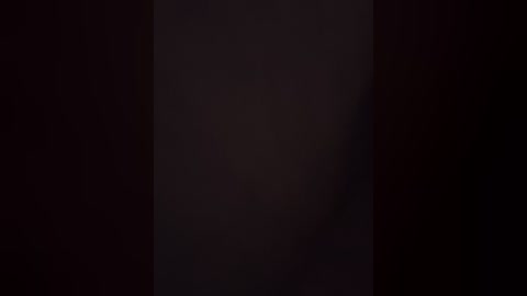 A dimly lit video showing a man's face partially obscured by a vertical dark shadow. The background is dark, with subtle variations in light and shadow. The image is grainy, giving it a vintage film effect.