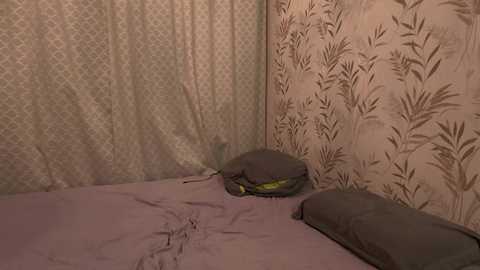 Video of a dimly lit, cramped bedroom corner with a small bed covered in a gray blanket, a pillow, and a green backpack. Wallpaper features a leaf pattern in brown and beige.