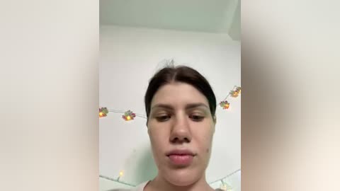 A video of a young woman with light skin, dark hair, and minimal makeup, standing in front of a white wall adorned with colorful string lights, taken from a low angle.