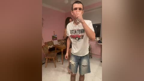 Video of a young man with short hair in a white \"Desert Town\" T-shirt and ripped jeans, drinking water in a pink room with a wooden table, chairs, and a pink cabinet.