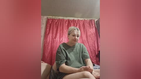 Video of a smiling, fair-skinned woman with short blonde hair, wearing a green t-shirt and shorts, sitting on a bed with red curtains and beige walls in the background.