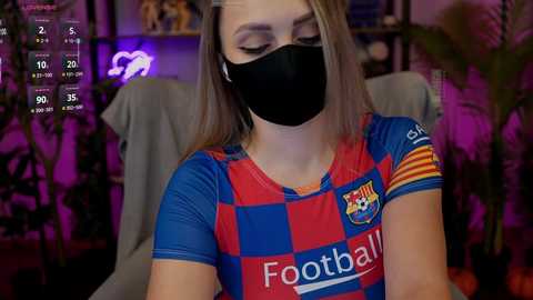 Video of a woman with light skin, wearing a black face mask, a blue and red Barcelona soccer jersey, and a yellow armband, seated indoors, with a digital clock and blurred background.