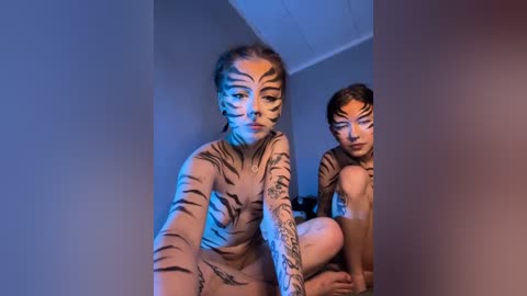 Video of a woman with tiger face paint, holding her legs, standing in front of a mirror, blue lighting, minimalist background, tattoos on her arms.