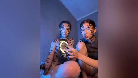 Video of two women with dark hair, tiger-striped makeup, and matching striped tops, sitting on a bed under blue lighting, holding a camera.