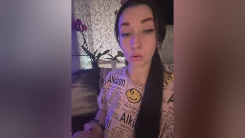 Video of a young woman with pale skin, dark hair, and thick eyebrows, wearing a \"Alaska Dispatch News\" shirt, sitting indoors with a dark-haired man partially visible. Background features a floral-patterned wallpaper and a purple orchid.