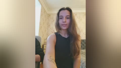 Video of a young woman with long, wavy brown hair in a black sleeveless top, standing in a dimly lit room with patterned wallpaper and a bed in the background.