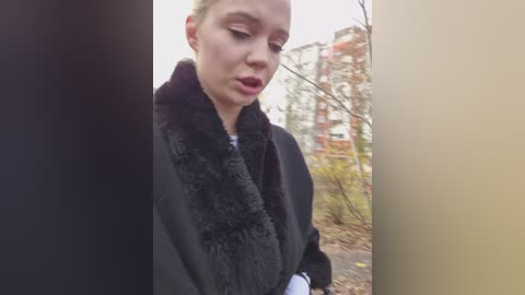 A video of a Caucasian woman with blonde hair, wearing a black coat and scarf, looking down thoughtfully in an outdoor park setting with urban buildings in the background.