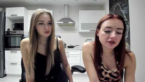 Video of two young women in a modern kitchen. One with long blonde hair in a black top, the other with red hair in a leopard-print top, both looking introspective.