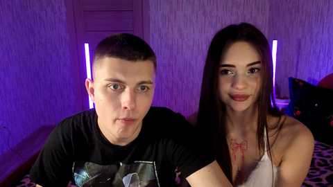 Video of a young couple with fair skin, short-cropped hair, and wearing casual T-shirts, sitting on a leopard-print couch against a purple wall. The woman has long dark hair and glasses, smiling, while the man looks serious.