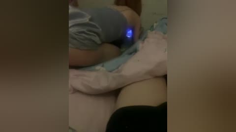 A blurry video showing a person lying on a bed, partially obscured by a person's legs and hands. The room has dim lighting, and the individual is wearing light-colored clothing.