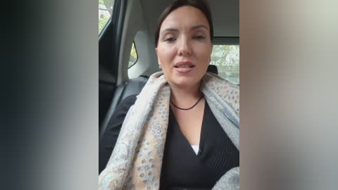 Video of a woman with light skin, brown hair, and light makeup, wearing a black V-neck top, gray patterned cardigan, and a black choker. She's seated in a car, looking relaxed, with blurred car window and greenery background.