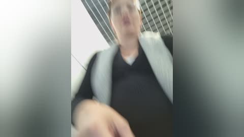 Video of a person in a black sweater with a white jacket, taken from a low angle, focusing on their torso and head, with a blurred background featuring a grid ceiling.