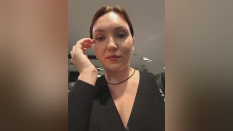 Video of a fair-skinned woman with dark brown hair, wearing a black top and a black choker, adjusting her hair. Background shows a modern, well-lit room with grey walls and recessed lighting.