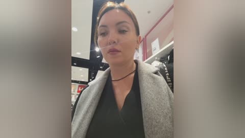 Video of a woman with light skin and brown hair, wearing a black top and gray coat, standing in a brightly lit, modern clothing store with shelves in the background.