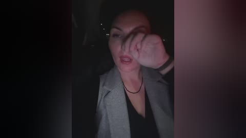 A dimly lit video of a woman with medium skin tone, dark hair, and a gray blazer, shielding her face with her hand, seated in a car at night.