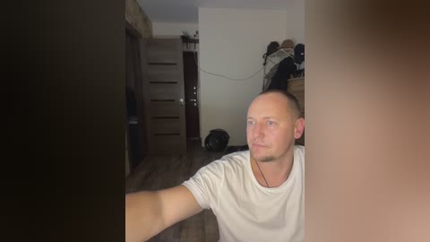 Video of a bald Caucasian man in a white t-shirt, sitting on a chair in a dimly lit room with wooden floors, a dark doorway, and cluttered background.