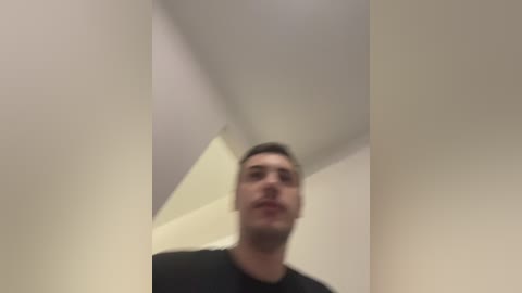 A video of a young man with short dark hair, wearing a black t-shirt, standing in a minimalist, modern room with white walls and a white ceiling.