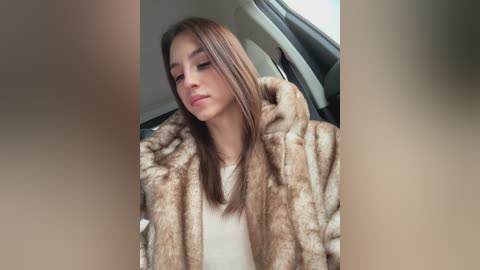 A video of a young woman with straight, light brown hair, wearing a luxurious brown fur coat, seated in a car, looking downward. The background features a car interior with neutral tones.