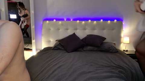 Video of a modern bedroom with a plush white tufted headboard, purple LED strip lighting, dark grey bedding, and two women in lingerie. One woman dances seductively in the doorway.