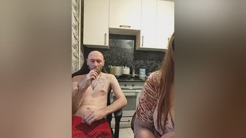 A video of a bald, shirtless man in red shorts seated at a kitchen table, holding a phone to his ear, with a woman in a textured blouse in the background.