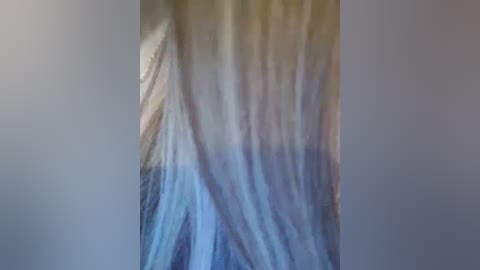 A close-up video of a person's shoulder and upper back, featuring long, straight, light brown hair with subtle highlights. The background is a blurred mix of muted beige and blue tones.
