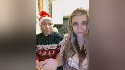 Video of an elderly man in a red Santa hat and a Nordic-patterned sweater, seated beside a young woman with long, blonde hair and a low-cut top, in a modern kitchen with white cabinets and gray backsplash.