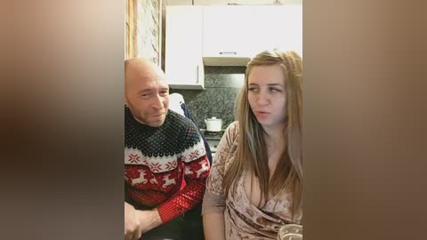 Video of a middle-aged bald man in a red and white sweater, and a young woman with long blonde hair in a beige blouse, sitting closely together in a modern kitchen with white cabinets and dark countertops.