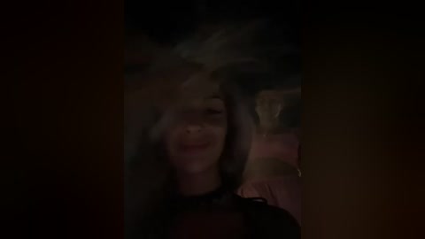 A dark, blurry video of a woman with long hair, smiling, with a smoke-filled background. She wears a black top. The image is dimly lit, giving it a mysterious, moody ambiance.