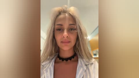 Video of a young woman with light skin, long blonde hair, wearing a white button-up shirt, black choker, and a neutral expression. The background is blurred, featuring a white ceiling and wooden furniture.