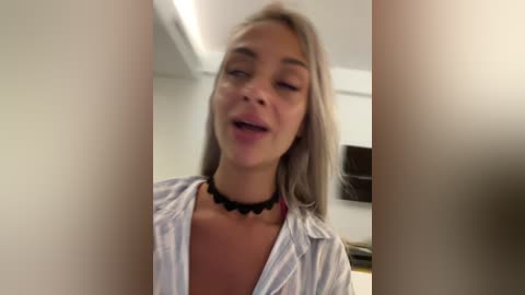 Video of a smiling, light-skinned woman with straight, shoulder-length blonde hair, wearing an unbuttoned white shirt and a black choker necklace. Background shows a white wall and a blurred, dark object.